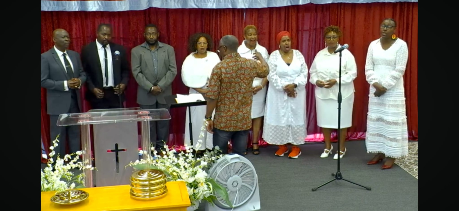 Patrick Mose directing a choir during a worship service, singers standing together in front of a red backdrop